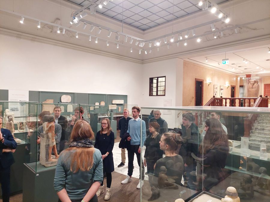 zur Vergrößerungsansicht des Bildes: Förderer und Stipendiaten stehen um den Vitrinen im Ägyptischen Museum und hören den Ausführungen der Gästeführerin zu