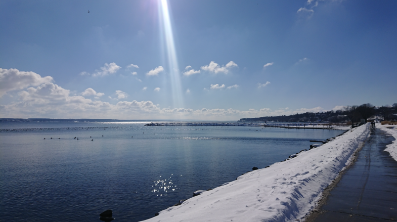 Ein blauer See befindet sich in der linken Bildhälfte. Der Himmel ist ebenfalls blau. Von der oberen Bildmitte scheint ein Sonnenstrahl über das Bild. Rechts ist ein zugeschneiter Weg zu sehen.