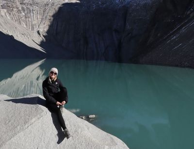 Person sitzt auf einem Stein, dahinter ein türkisfarbener See und hohe Felsen bei Sonnenschein