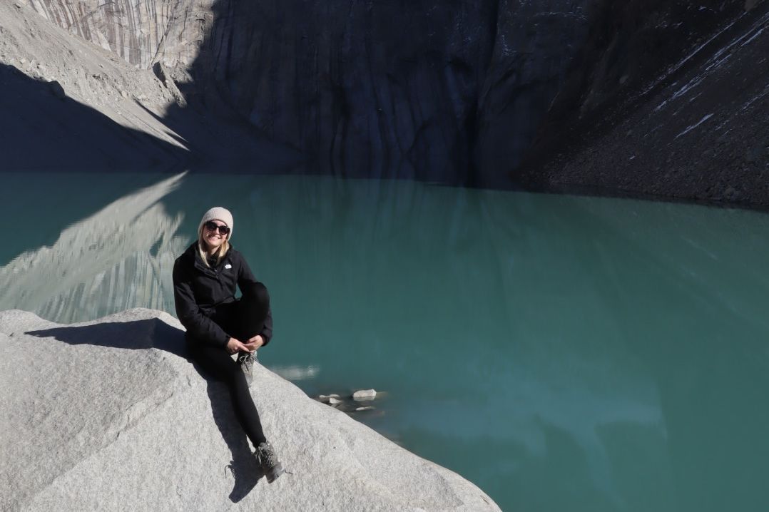 enlarge the image: Person sitzt auf einem Stein, dahinter ein türkisfarbener See und hohe Felsen bei Sonnenschein