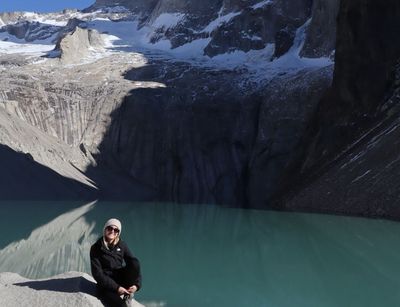 Person sitzt auf einem Stein, dahinter ein türkisfarbener See und hohe Felsen bei Sonnenschein