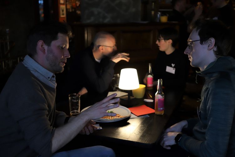 Two pairs of people sitting at the table in a dark cafe and talking to each other. There is a lamp, drinks and a plate with pizza on the table.