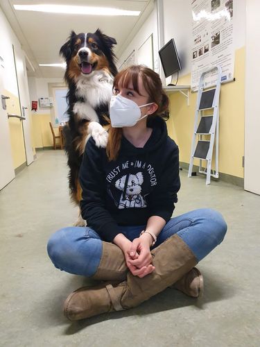 Louise sitzt in der Bildmitte im Schneidersitz auf dem Boden. Ein Hund hockt hinter ihr. Er hat eine Pfote auf ihre linke Schulter gelegt.