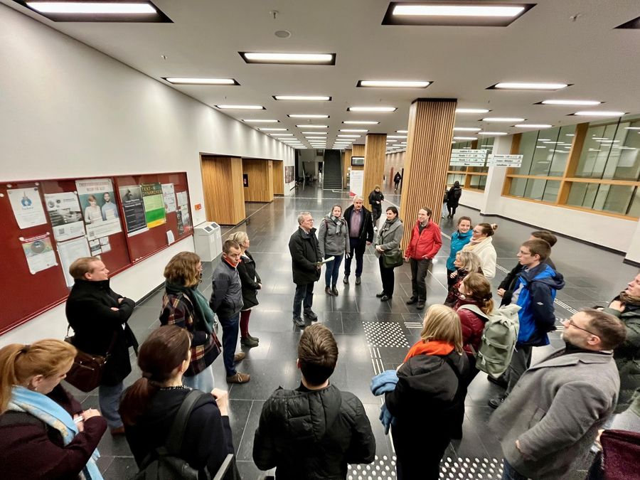 zur Vergrößerungsansicht des Bildes: die Gäste der Führung durchs Bibliotop stehen zu Beginn im Hörsaalgebäude, sie stehen im Kreis um Professor Lokatis und hören seinen Ausführungen zu