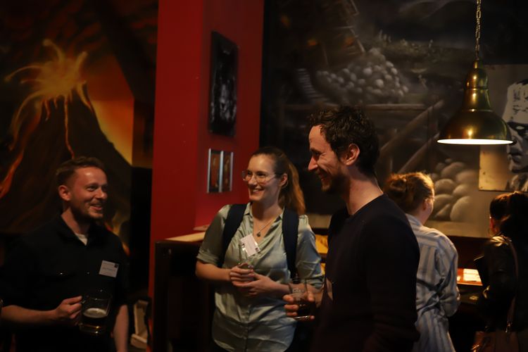 Two men and a woman standing in circle in a cafe, holding drinks and smiling.