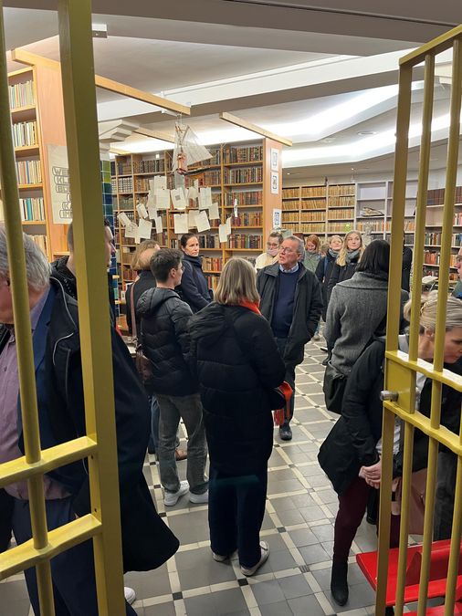 zur Vergrößerungsansicht des Bildes: Förderer und Stipendiaten stehen im Bibliotop und hören den Ausführungen von Professor Lokatis, der in der Mitte zu steht, zu, im Vordergrund ist der vergitterte Eingang zum ehemaligen Tresorraum zu sehenen