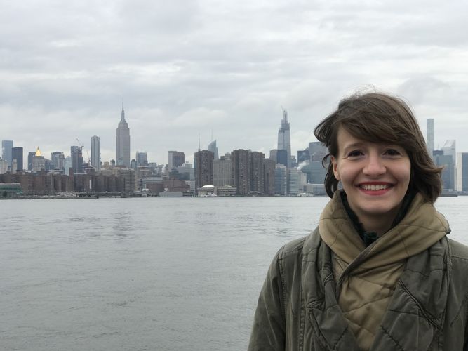 Eine Frau lacht in die Kamera. Sie steht am rechten Rand des Bildes. Im Hintergrund ist die Skyline von Manhatten, New York City, zu sehen.