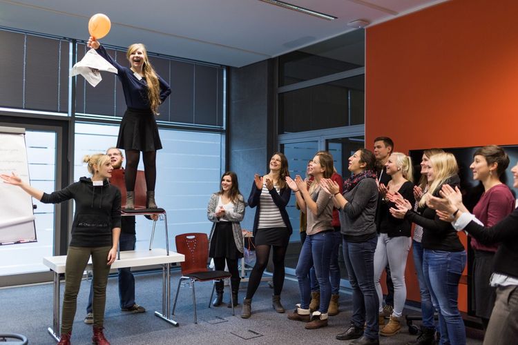 Seminarraum: Eine Person steht auf einen Stuhl, der auf einen Tisch steht und hält einen orangenen Luftballon in die Höhe. Sie blickt freudig auf ihn. Rechts im Bild steht eine Gruppe Studierender, die zu ihr blicken und applaudieren, Foto: Christian Hüller