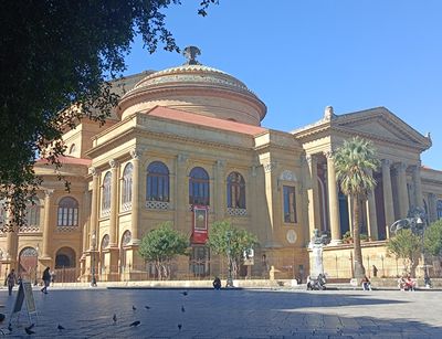 Sandsteinfarbenes Theater mit Kuppel und Säulen, davor ein Platz, Bei Sonnenschein