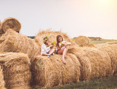 Man sieht zwei Mädchen mit Blumen in den Haaren auf Heuballen sitzen. Im Hintergrund steht die Sonne tief.