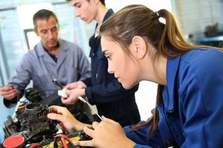 Farbfoto: Im Vordergrund steht eine junge Frau im blauen Arbeitskittel mit einem Maulschlüssel in der Hand und arbeitet an einem mechanischen Werkstück. Im Hintergrund steht ein älterer Mann, der einem sehr jungen Mann etwas an einem anderen mechanischem Teil erklärt.