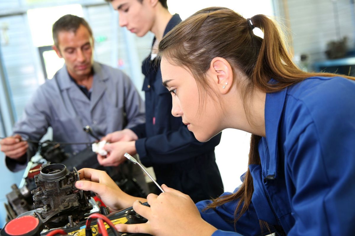 zur Vergrößerungsansicht des Bildes: Farbfoto: Im Vordergrund steht eine junge Frau im blauen Arbeitskittel mit einem Maulschlüssel in der Hand und arbeitet an einem mechanischen Werkstück. Im Hintergrund steht ein älterer Mann, der einem sehr jungen Mann etwas an einem anderen mechanischem Teil erklärt.