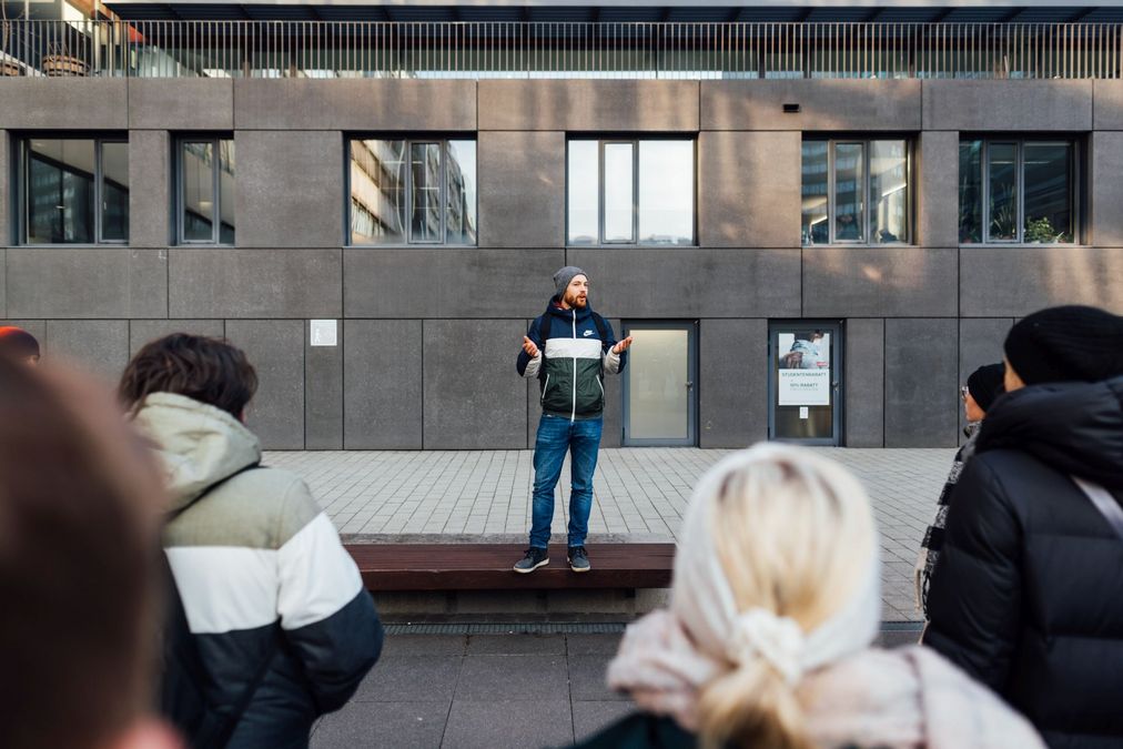 zur Vergrößerungsansicht des Bildes: Campusguide steht auf einer Bank im Uni-Innenhof und im Vordergrund sieht man Teilnehmende an der Campustour, Foto: Christian Hüller