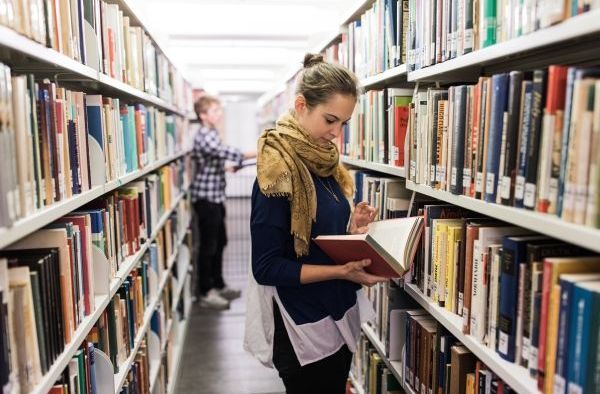 Eine Frau steht in einer Bibliothek zwischen zwei Bücherregalen und liest ein Buch