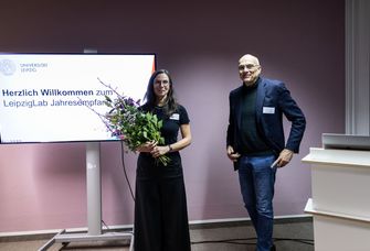 Zwei Personen, eine Frau mit Blumenstrauß und ein Mann, stehen vor Bildschirm mit Aufschrift