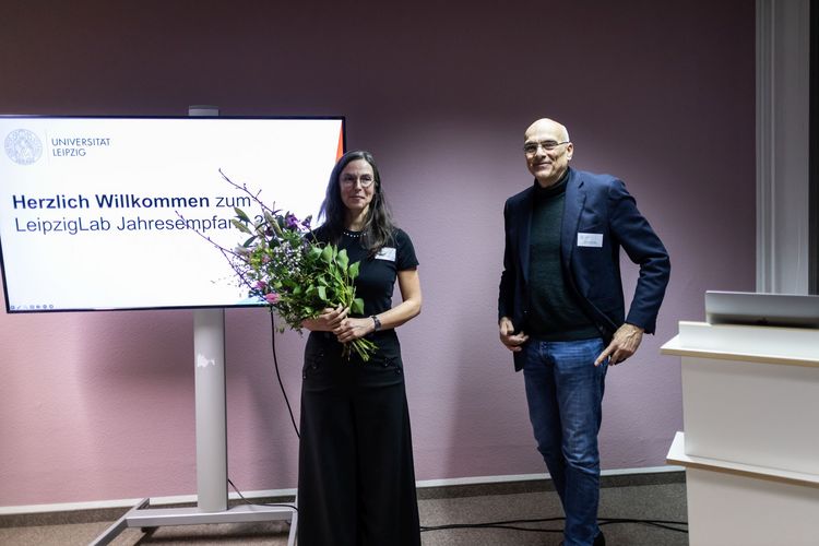 Zwei Personen, eine Frau mit Blumenstrauß und ein Mann, stehen vor Bildschirm mit Aufschrift