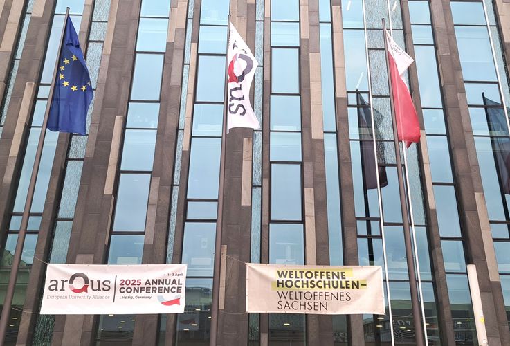 Zu sehen sind die Fahnen der EU, von Arqus und der Universität Leipzig, die vor einem Gebäue wehen. Darunter hängt ein Banner mit dem Hinweis auf die Jahreskonferenz und ein Banner auf dem "Weltoffene Hochschulen - Weltoffenes Sachsen" steht.