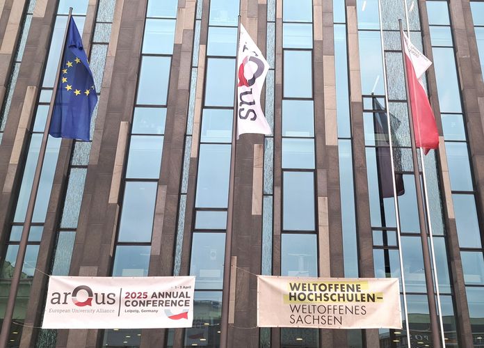 Zu sehen sind die Fahnen der EU, von Arqus und der Universität Leipzig, die vor einem Gebäue wehen. Darunter hängt ein Banner mit dem Hinweis auf die Jahreskonferenz und ein Banner auf dem "Weltoffene Hochschulen - Weltoffenes Sachsen" steht.