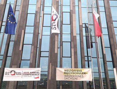 Zu sehen sind die Fahnen der EU, von Arqus und der Universität Leipzig, die vor einem Gebäue wehen. Darunter hängt ein Banner mit dem Hinweis auf die Jahreskonferenz und ein Banner auf dem "Weltoffene Hochschulen - Weltoffenes Sachsen" steht.