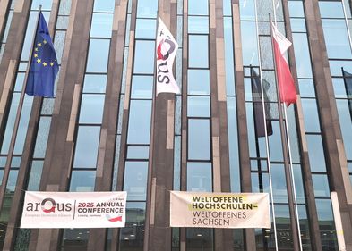 Zu sehen sind die Fahnen der EU, von Arqus und der Universität Leipzig, die vor einem Gebäue wehen. Darunter hängt ein Banner mit dem Hinweis auf die Jahreskonferenz und ein Banner auf dem "Weltoffene Hochschulen - Weltoffenes Sachsen" steht.