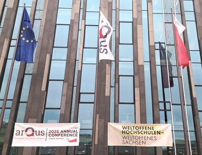 Zu sehen sind die Fahnen der EU, von Arqus und der Universität Leipzig, die vor einem Gebäue wehen. Darunter hängt ein Banner mit dem Hinweis auf die Jahreskonferenz und ein Banner auf dem "Weltoffene Hochschulen - Weltoffenes Sachsen" steht.