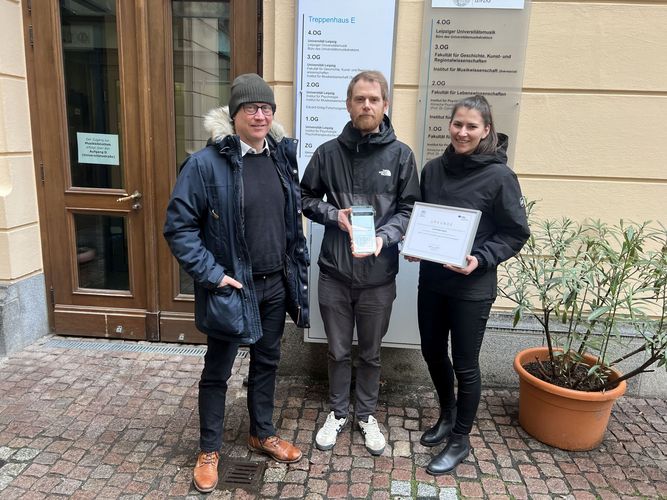 Auf dem Bild sind Johanna Kaiser und Prof. Dr. Julian Schmitz (Mitte) sowie VBG-Mitarbeiter Martin Kirsten (links) zu sehen. Kaiser hält eine Urkunde hoch, Schmitz einen gläsernen Blitz (Preis).hnet. 