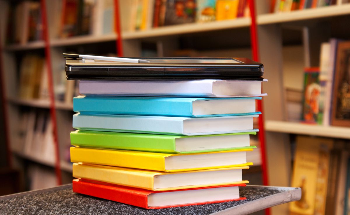 zur Vergrößerungsansicht des Bildes: Zu sehen ist ein Stapel bunter Bücher mit einem Tablet. Sie liegen auf einem Tisch in einer Bibliothek