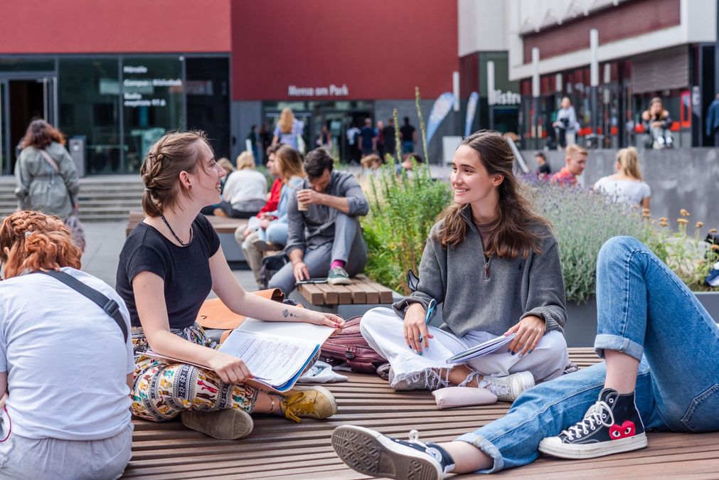 enlarge the image: Zwei Studentinnen sitzen auf einer Bank im Leibnizforum und sprechen miteinander, um sie herum herrscht reges Treiben im Uni-Innenhof, Foto: Kirsten Nijhof