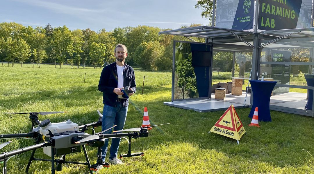 Auf dem Bild ist Wirtschaftsinformatiker Martin Schieck auf einer Wiese neben einer Drohne zu sehen. Neben ihm das Smart Farming Lab.ersuchsfeld. Foto: Diana Smikalla