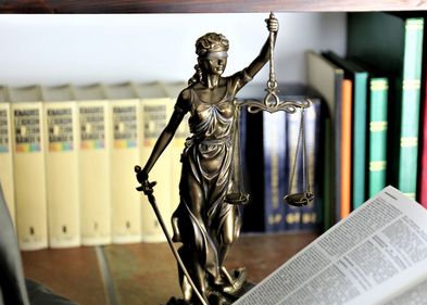 Farbfoto: eine kleine bronzefarbene Statue der Justizia mit erhobenem Schwert, verbundenen Augen und Waagschale steht auf einem Tisch. Im Vordergrund liegt ein aufgeschlagenes Buch mit Gesetzestexten. Im Hintergrund stehen Bücher in einem Regal.