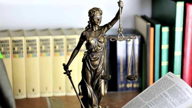 Farbfoto: eine kleine bronzefarbene Statue der Justizia mit erhobenem Schwert, verbundenen Augen und Waagschale steht auf einem Tisch. Im Vordergrund liegt ein aufgeschlagenes Buch mit Gesetzestexten. Im Hintergrund stehen Bücher in einem Regal.