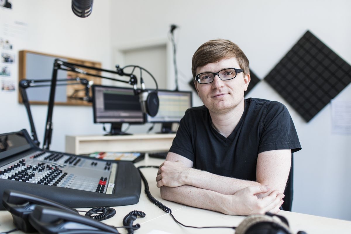 zur Vergrößerungsansicht des Bildes: Foto: ein Mann in schwarzem T-Shirt gekleidet sitzt in einem Studio vor einem Mischpult 