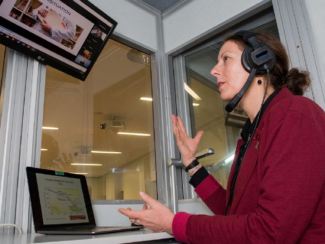 Eine Frau sitzt in einer Dolmetschkabine, gestikuliert mit den Händen und blickt auf einen Monitor mit einer Zoom-Konferenz. Foto: Bodo Tiedemann