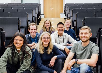 Farbfotografie von sieben Studierenden, den Studienbotschafterinnen, die im Gang, zwischen den Stuhlreihen eines Hörsaals auf dem Boden sitzen