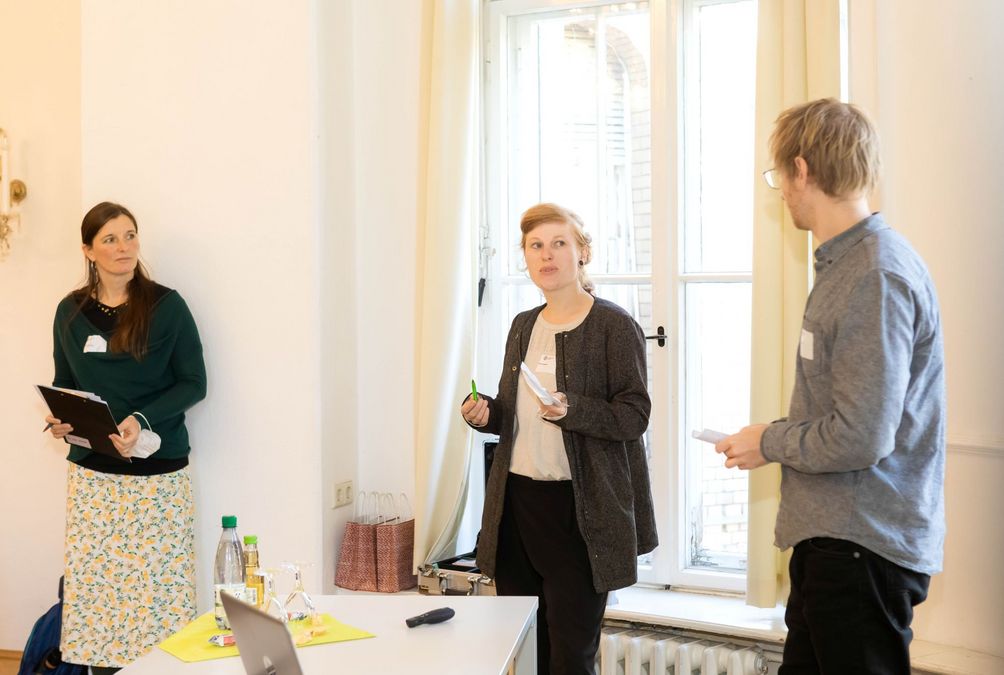 zur Vergrößerungsansicht des Bildes: Zwei Menschen stehen vor dem Publikum und beantworten Fragen. Daneben steht beobachtend Programmkoordinatorin Dr. Nicole Koburger. 