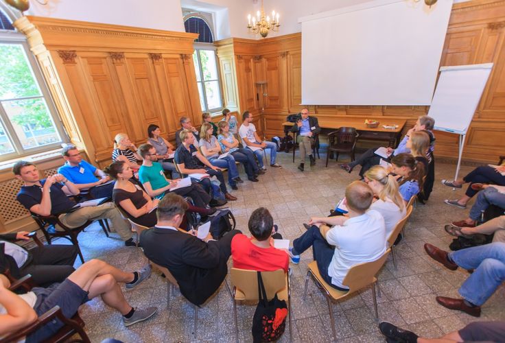 Die Fotografie zeigt eine Gruppe von ca. 30 Personen, die in einem Stuhlkreis zusammensitzen. Eine Person spricht und die anderen hören zu und machen sich Notizen.