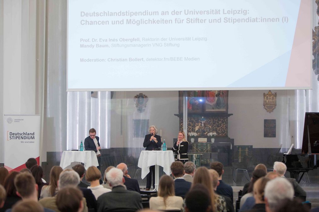 zur Vergrößerungsansicht des Bildes: Rektorin, Mandy Baum von VNG und Moderator Christian Bollert stehen auf der Bühne und sprechen zu den Gästen der Stipendienfeier, im Hintergrund ist der Altar im Paulinum zu sehen