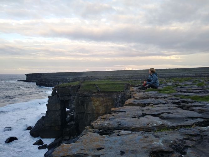 Eine Person sitzt auf einer begrasten Klippe in der rechten Bildhälfte. In der linken Bildhälfte ist der Atlantik zu sehen.