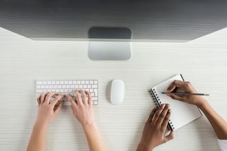 Blick von oben auf einen Computerbildschirm mit Tastatur und einen Notizblock. Am unteren Bildrand erscheinen Arme, die die Tastatur bedienen und auf dem Block schreiben.