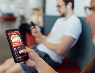 Studentin hält ein Smartphone in den Händen, wo eine Folge vom Podcast Auf einen Kaffee mit auf dem Display zu sehen ist, im Hintergrund sitzt ein Student, Foto: Christian Hüller