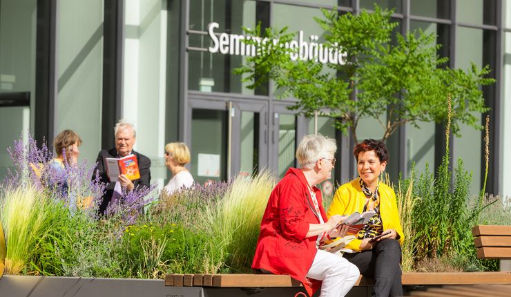 Farbaufnahme: zwei ältere Frauen sitzen im Uni-Innenhof und unterhalten sich.