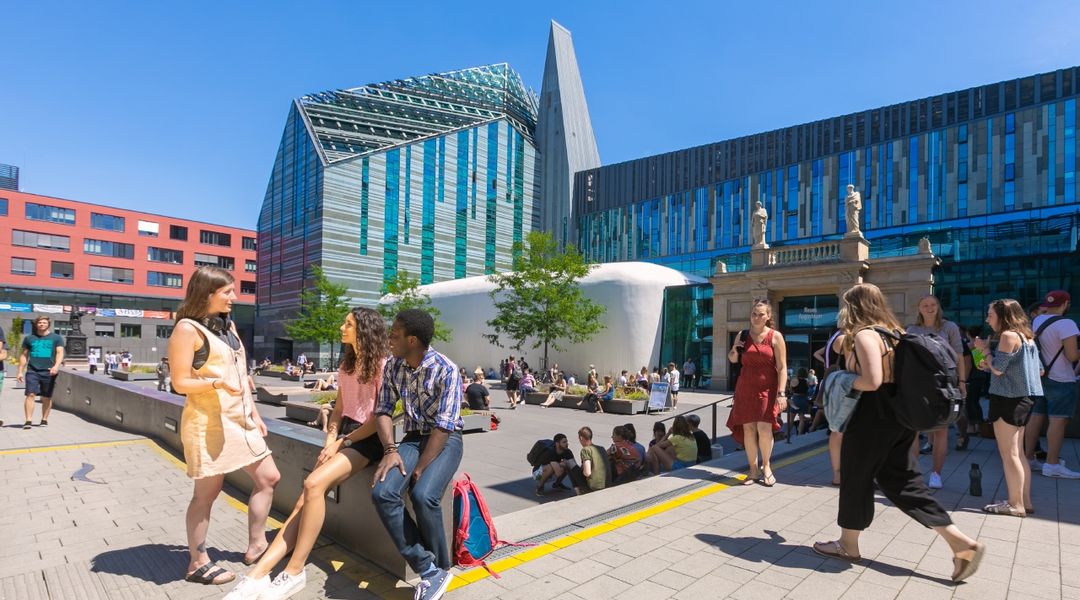 Farbfotografie vom belebten Innenhof des Campus Augustusplatz in sommerlicher Atmosphäre. Im Fokus des Bildes steht eine Gruppe von drei jungen Menschen, die sich unterhalten. Zwei von Ihnen sitzen auf einer halbhohen Mauer. Eine weitere junge Frau steht ihnen zugewandt und spricht mit ihnen. Im Hintergrund sieht man mehrere kleine Gruppen von Menschen, die auf dem Campus verweilen oder über den Campus laufen.