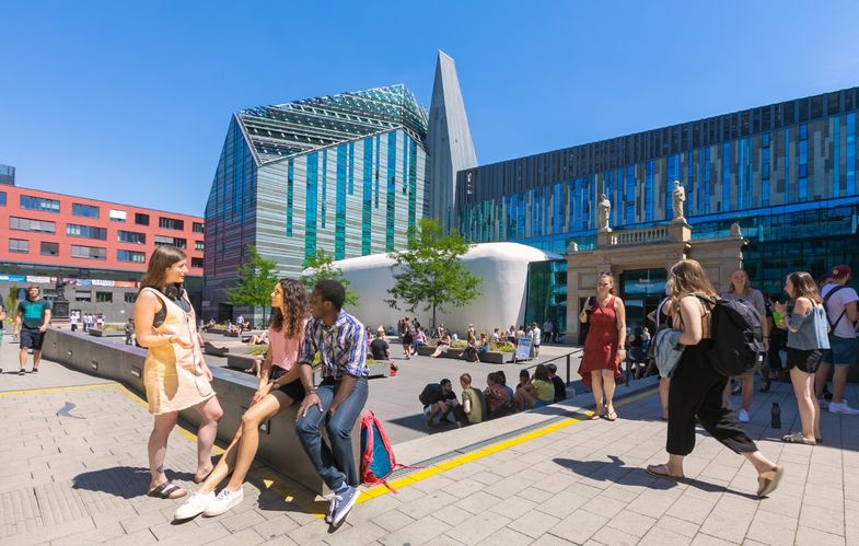 Farbfotografie vom belebten Innenhof des Campus Augustusplatz in sommerlicher Atmosphäre. Im Fokus des Bildes steht eine Gruppe von drei jungen Menschen, die sich unterhalten. Zwei von Ihnen sitzen auf einer halbhohen Mauer. Eine weitere junge Frau steht ihnen zugewandt und spricht mit ihnen. Im Hintergrund sieht man mehrere kleine Gruppen von Menschen, die auf dem Campus verweilen oder über den Campus laufen.