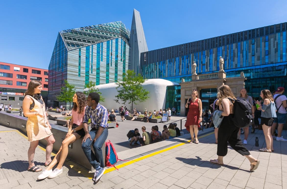 zur Vergrößerungsansicht des Bildes: Farbfotografie vom belebten Innenhof des Campus Augustusplatz in sommerlicher Atmosphäre. Im Fokus des Bildes steht eine Gruppe von drei jungen Menschen, die sich unterhalten. Zwei von Ihnen sitzen auf einer halbhohen Mauer. Eine weitere junge Frau steht ihnen zugewandt und spricht mit ihnen. Im Hintergrund sieht man mehrere kleine Gruppen von Menschen, die auf dem Campus verweilen oder über den Campus laufen.