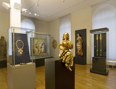 Foto: Ausstellungsstücke aus der Kustodie werden gezeigt, im Vordergrund eine goldene Statue, im Hintergrund weitere religiöse Statuen und Malereien.