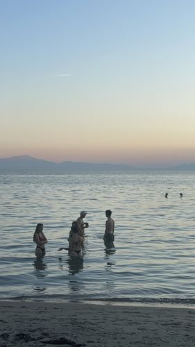 Vier Personen stehen kniehoch im Wasser am Strand, im Hintergrund geht in dämmerndem Licht die Sonne unter. 