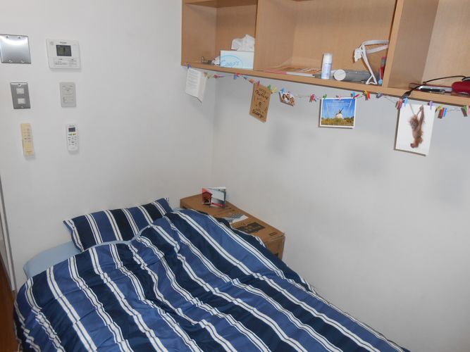 Zu sehen ist ein Bett, mit einer dunkelblau und hellblau gestreifter Bettwäsche darauf. Über dem Bett hängt ein braunes Regal, welches mit Postkarten geschmückt ist.