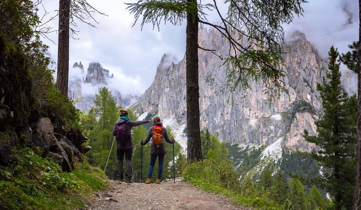 Zwei junge Frauen in Wanderkleidung befinden sich in einer Berglandschaft. Ihr Blick ist in Richtung des Berggipfels gerichtet. Eine der Frau deutet mit ihrem Finger in Richtung Gipfel.
