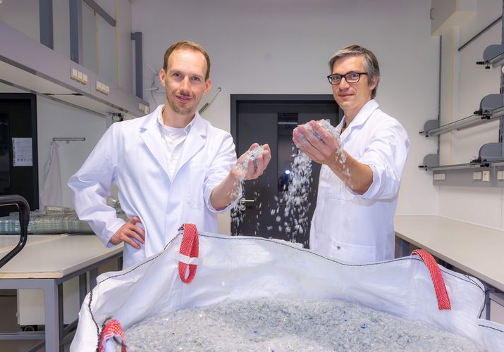 Dr. Ronny Frank und Dr. Christian Sonnendecker stehen vor einem Sack voll kleingehäckselten Plastikteilen. 