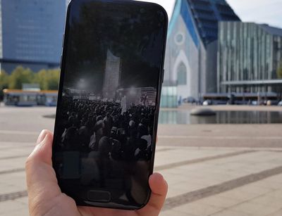 Farbfoto: Im Mittelpunkt ist ein Handy zu sehen, welches ein historisches schwarz-weiß Bild von einer Montagsdemonstration im Herbst 1989 zeigt. Das Handy wird von einer Hand gehalten. Im Hintergrund ist der Campus Augustusplatz mit den neuen Gebäuden der Universität Leipzig "Paulinum" und "Augusteum" zu sehen.